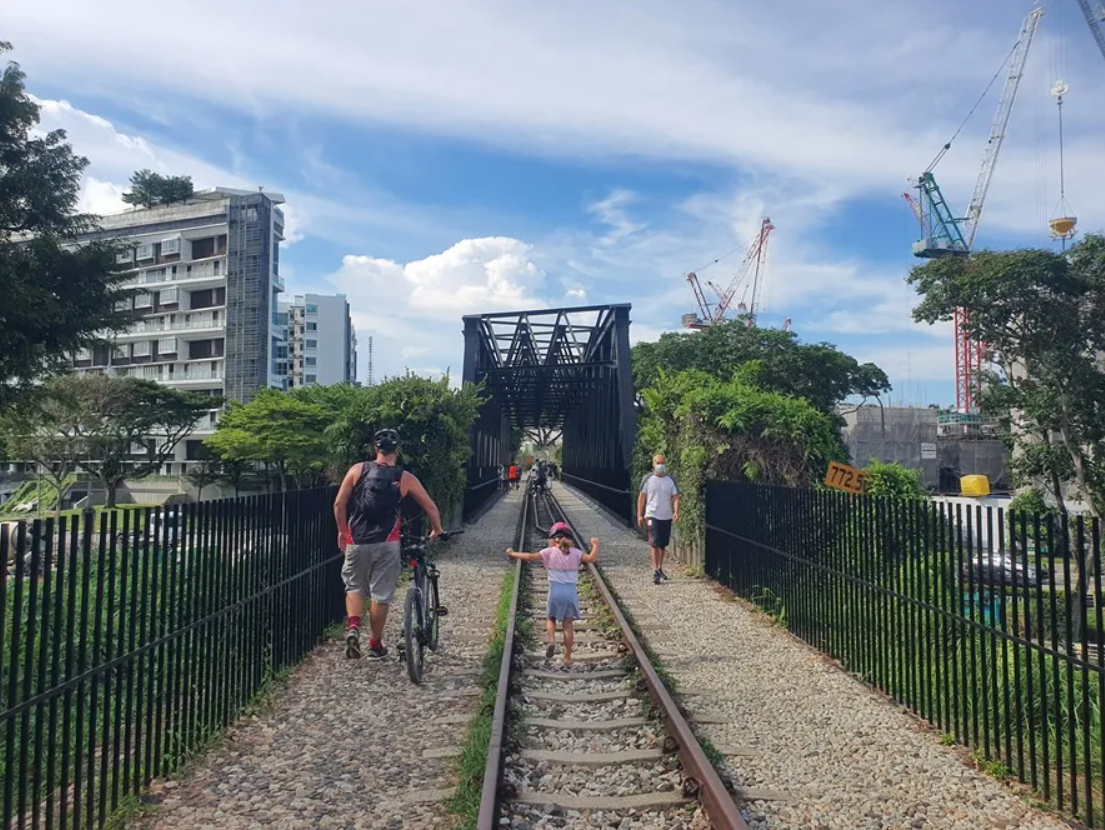 People are walking along a gravel path beside a railway track, heading towards a steel bridge, with buildings and cranes in the background.